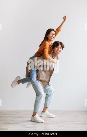 Bel giovane uomo asiatico che dà cavalcata piggyback alla sua ragazza contro la parete bianca dello studio, ritratto a tutta lunghezza Foto Stock