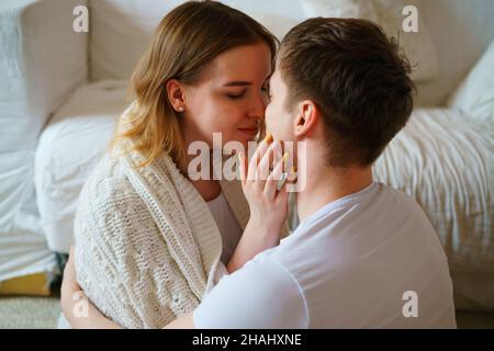 Felice coppia sposata di proprietari di case millennial girato ritratto. Marito e moglie sono seduti sul pavimento vicino al divano in un abbraccio in una coperta, baciare, abbracciare con amore, rilassarsi in una nuova casa accogliente Foto Stock