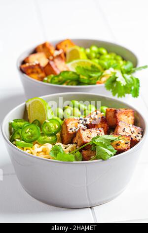 Zuppa di ramen di tagliatelle vegane con tofu marinato, fagioli edamame e peperoni caldi in una ciotola grigia su sfondo bianco, primo piano. Foto Stock