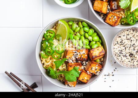 Zuppa di ramen di tagliatelle vegane con tofu marinato, fagioli edamame e peperoni caldi in una ciotola grigia su sfondo bianco piastrellato, vista dall'alto. Foto Stock