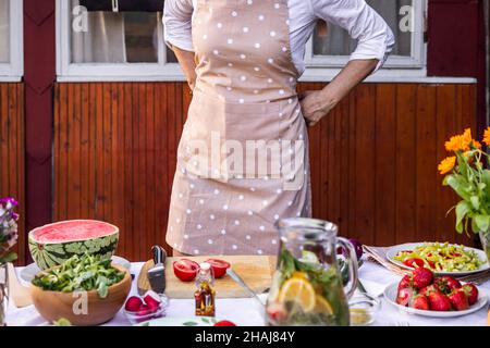 Donna che lega grembiule e si prepara per cucinare. Preparazione di cibi freschi e sani per feste in giardino all'aperto Foto Stock