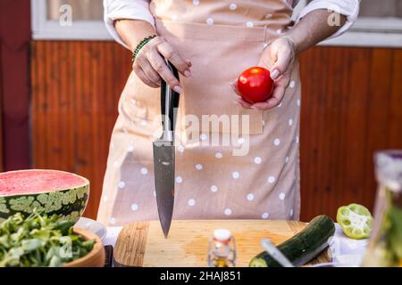 Donna con grembiule e coltello da cucina e pomodoro rosso. Insalata di verdure da cucina Foto Stock