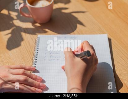 Donna mano scrittura piani in notebook o pianificatore su legno scrivania con luce solare e tazza di caffè. Foto Stock