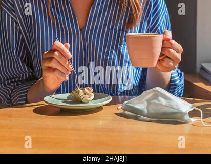 Maschera medica sul tavolo da caffè mentre la persona ha la colazione, mangiare dolce e bere caffè. Foto Stock