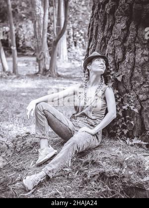 Una donna in tute e cappello in denim siede sotto un albero di foresta appoggiata sul suo tronco e guarda in su da sogno. Foto Stock