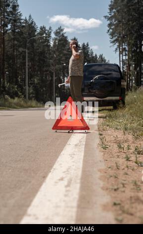 Segnale di attenzione triangolare sulla strada dopo un guasto all'auto e ma che parla sullo smartphone, in attesa di un intervento di riparazione. Foto Stock