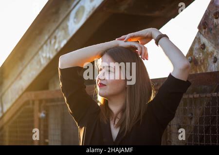 Ritratto di bella giovane donna in città al tramonto. Godimento di una giornata di sole. Donna bruna alla moda Foto Stock