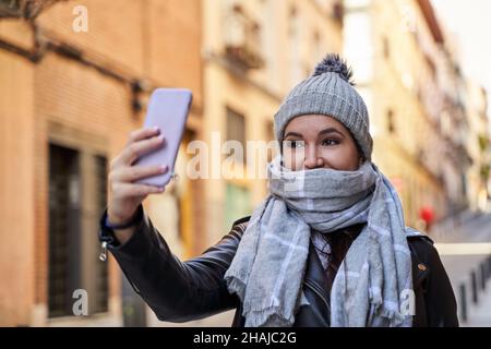 Giovane uomo in berretto e sciarpa sorridente prendendo un selfie via smartphone in una giornata fredda sulla strada della città. Foto Stock