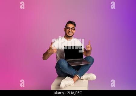 Giovane ragazzo arabo che tiene un laptop con schermo vuoto, mostrando il pollice in su, consigliando il nuovo sito web in luce al neon. Mockup Foto Stock