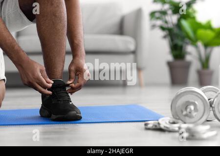 L'irriconoscibile atleta maschile nero da millennial si unisce alle sneakers sul tappetino interno della stanza con manubri Foto Stock