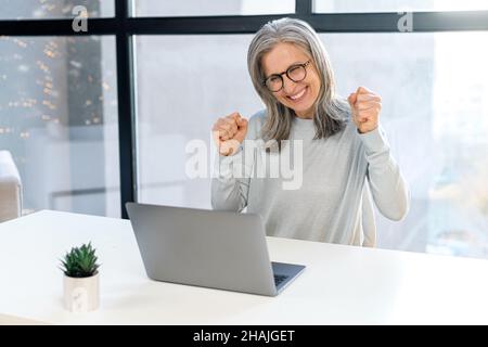 Obiettivo raggiunto. Esilarante sorridente matura donna dai capelli grigi urla sì e alzando le mani in un gesto di vittoria, seduto davanti al portatile in ufficio e gioire per vincere Foto Stock