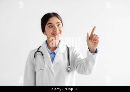 Sorridendo piuttosto giovane medico indù donna in cappotto bianco con stetoscopio punta il dito a spazio vuoto Foto Stock