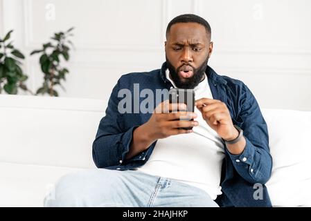 Scioccato giovane afro-americano barbed ragazzo in casual fissando lo schermo dello smartphone seduto sul divano in appartamento moderno, deluso ragazzo nero ricevuto inaspettato cattivo nuovo Foto Stock