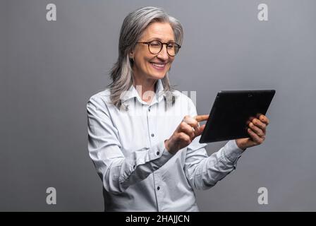 Donna anziana moderna in abbigliamento formale utilizzando tablet digitale isolato su grigio, ritratto di una donna matura ufficio utilizzando la tecnologia online per fare affari, app computer per la contabilità Foto Stock