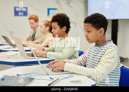 Vista laterale di diversi gruppi di bambini che utilizzano computer portatili alla scrivania in un'aula moderna, spazio per le copie Foto Stock