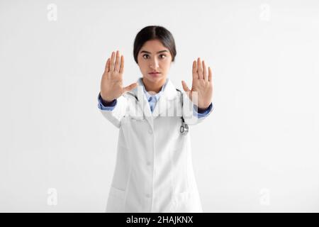 Serio giovane medico indiano signora in mano con stetoscopio fa stop segno con le mani su sfondo bianco Foto Stock