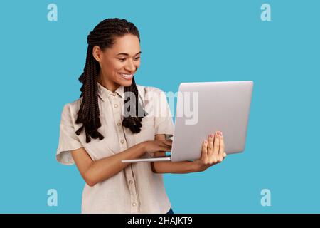 Donna sorridente con lucchetti neri che tengono un computer portatile, ride mentre guarda video divertenti dai social network, naviga in internet, indossa una camicia bianca. Studio interno girato isolato su sfondo blu. Foto Stock