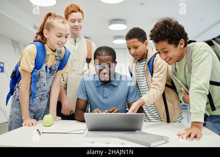 Gruppo di bambini diversi che si levano in piedi intorno al giovane insegnante afro-americano usando il laptop in classe Foto Stock