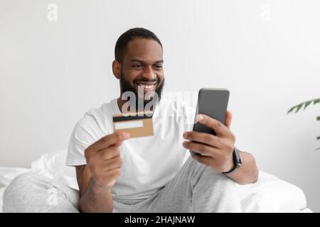 Sorridente nero barbuto maschio su letto bianco in camera da letto fa acquisti online con il telefono Foto Stock