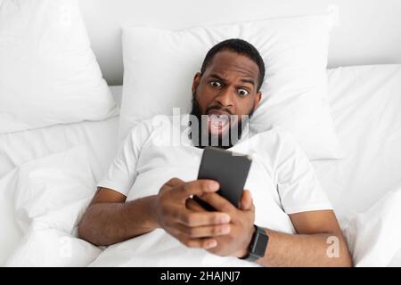 Sorpreso allegro uomo americano africano millenario con barba e bocca aperta giace su letto bianco Foto Stock