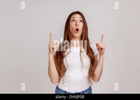 Donna sorpresa con capelli scuri che puntano fino a spazio vuoto copia sopra la testa e guardando con stupore in su, mostrando parete vuota, indossando T-shirt bianca. Studio interno girato isolato su sfondo grigio. Foto Stock