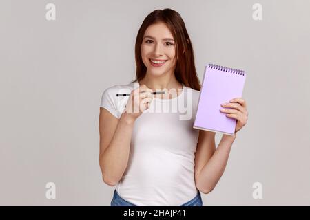 Positivo sorridente donna felice che punta a carta notebook con spazio vuoto per il testo commerciale, area pubblicitaria, indossare T-shirt bianco. Studio interno girato isolato su sfondo grigio. Foto Stock