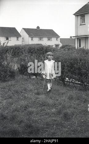 1960s, storica, all'esterno di una proprietà immobiliare, una giovane ragazza in piedi su un pezzo chiuso di prato, che tiene su due bastoni da passeggio, la sua gamba destra in un ferro di gamba di metallo, con reggette di cuoio, Inghilterra, Regno Unito. La polio e i ricci erano due condizioni che hanno causato la deformazione delle ossa delle gambe dei bambini e il ferro delle gambe o il sostegno delle gambe era un dispositivo che era stato usato dal 1940s per incoraggiare le ossa a crescere correttamente. Foto Stock