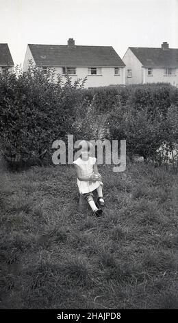 1960s, storica, all'esterno di una proprietà immobiliare, una giovane ragazza seduta su un pezzo chiuso di prato, la sua gamba destra in un ferro di gamba di metallo, con reggette in pelle, Inghilterra, Regno Unito. La polio e i ricci erano due condizioni che hanno causato la deformazione delle ossa delle gambe dei bambini e il ferro o il sostegno delle gambe era un dispositivo che era stato usato dal 1940s per incoraggiare le ossa a crescere correttamente. Foto Stock