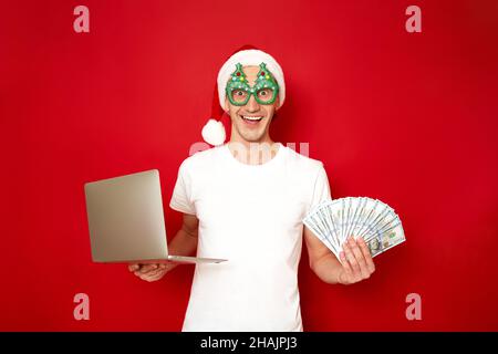 L'uomo eccitato in cappello e occhiali di nuovo anno alberi di Natale tiene il portatile e il ventilatore di soldi nelle sue mani Foto Stock