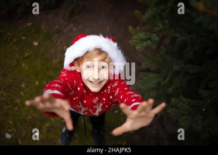 Un ragazzo in un maglione di Natale a maglia rossa con una renna di Natale e un cappello Babbo Natale nel parco allunga le mani dal basso fino alla camma Foto Stock