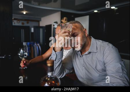 Bell'uomo europeo di mezza età in tuta informale che beve alcol, seduto su un bancone bar vicino alla sua ragazza a casa Foto Stock