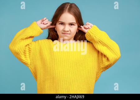 Ritratto di bambina in piedi con le orecchie chiuse per non sentire il consiglio dei genitori, ignorando l'educazione, indossando il maglione giallo casual stile. Studio interno girato isolato su sfondo blu. Foto Stock