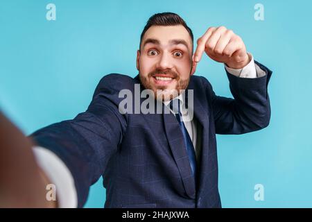 Uomo con un sorriso toothy in vestito, facendo selfie o videochiamate, guardando la telecamera POV, punto di vista della foto, puntando il dito verso il basso, iscriviti. Studio interno girato isolato su sfondo blu. Foto Stock