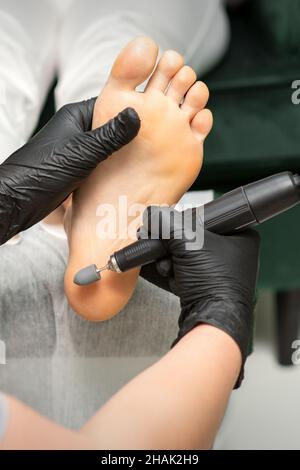 Peeling ai piedi femminili. Mani di podiatry specialista in guanti neri utilizzando speciale attrezzatura di macinazione lucidare il piede femminile da callo e mais nel salone di bellezza Foto Stock