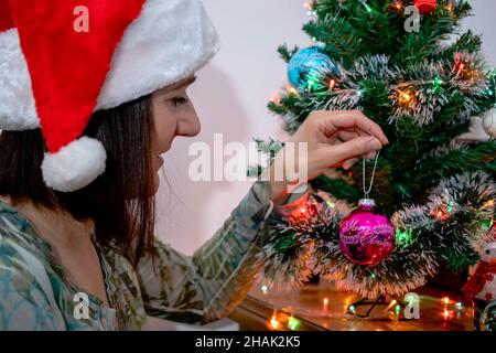 Donna che indossa un cappello di Babbo Natale, decorando un albero di Natale con un ornamento festivo rosa d'epoca con un segno di buon Natale. Vacanze invernali. Foto Stock