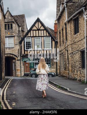 Donna che cammina lungo una strada a Burford, una città sul fiume Windrush, nelle colline del Cotswold, nel distretto di Oxfordshire occidentale dell'Oxfordshire, Inghilterra. Foto Stock