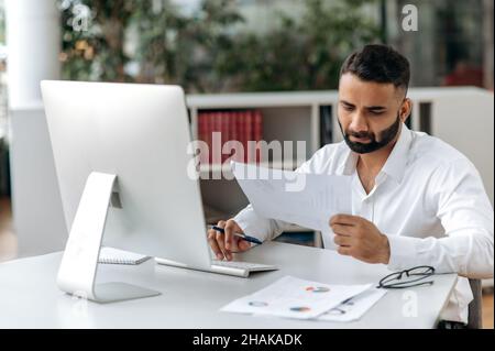 Occupato leader, manager o impiegato indiano di successo con barba, seduta in ufficio, uso del computer, studio di documenti e diagrammi, analisi, prendere appunti Foto Stock