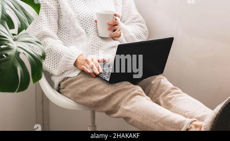 Una donna di mezza età si siede in una sedia con una tazza di tè o caffè e funziona su un computer portatile. Lavora online a casa Foto Stock