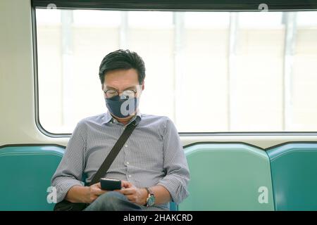 Uomo asiatico penduter indossare maschera viso guardando il suo smartphone, seduto all'interno di un treno in movimento metropolitana. Foto Stock