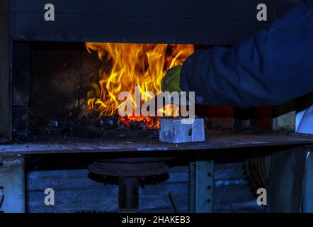 Il fabbro riscalda il metallo alla giusta temperatura prima di forgiarlo manualmente nel coltello da caccia perfetto. Bokeh. Foto Stock