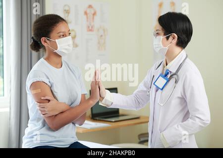 Adolescente ragazza in maschera medica dando cinque alti al medico dopo aver ottenuto un colpo di vaccino contro il coronavirus Foto Stock