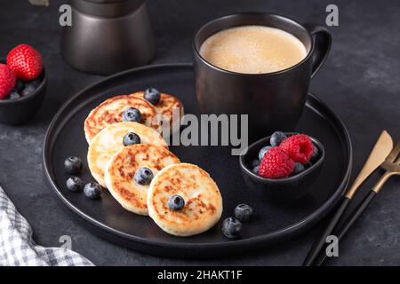 Pancake al formaggio fresco con lamponi freschi e mirtilli su piastra di ceramica scura. Tazza di caffè e macchina per il caffè Foto Stock