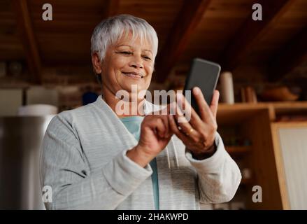 Felice multiculturale anziana femmina digitando un messaggio sullo smartphone mentre si trova in cucina moderna tenendo il caffè del mattino. Foto Stock