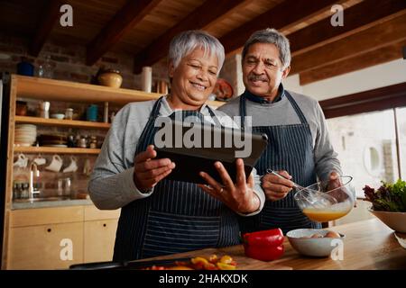 Basso angolo di Multi-culturale anziano coppia sorridente, utilizzando il tablet per la ricerca ricetta in cucina moderna. Foto Stock