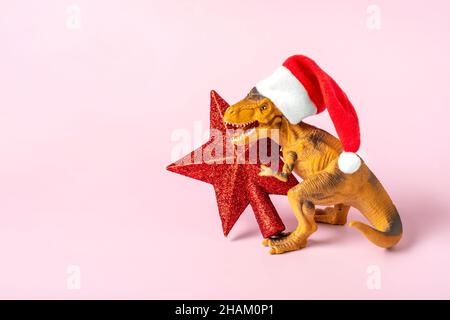 Il Rex del dinosauro in cappello rosso di Babbo Natale tiene la stella nelle relative zampe su sfondo rosa la vigilia di Capodanno o la vigilia di Natale scheda di festa d'arte creativa idea per Merry Foto Stock