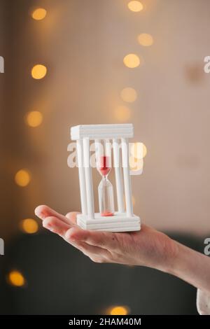 Le mani tengono una clessidra bianca sullo sfondo di una ghirlanda natalizia. Close-up clessidra. Il tempo è in esecuzione. Sabbia rossa in ore. L'anno nuovo è presto. Foto Stock