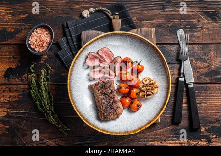 Filetto di agnello arrosto in piatto con pomodoro alla griglia e aglio, filetto di controfiletto di montone. Sfondo scuro. Vista dall'alto Foto Stock