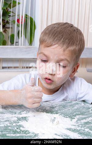 Un ragazzino emozionale e carino macchia il naso con farina e ti guarda. Gioca con la farina. Il bambino aiuta la madre con biscotti da forno, pizza a casa. Foto Stock