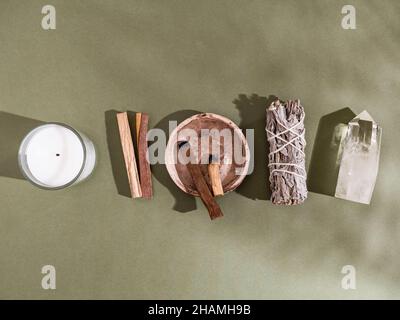 Articoli da collezione per la pulizia spirituale - pacchetto salvia, bastoni di palo santo incenso, candela e cristallo di quarzo su sfondo verde con ombra di piante. Da a. Foto Stock