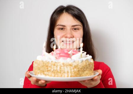 Una donna brunetta in abiti rossi tiene una grande torta bianca con candele, fa un desiderio Foto Stock